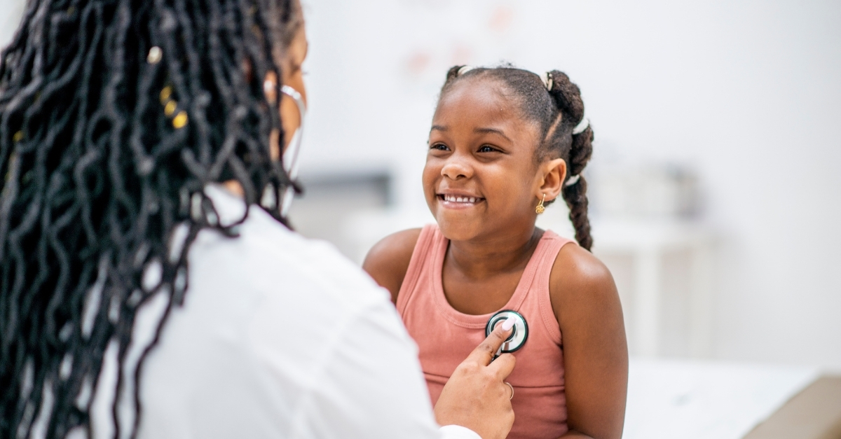 A smiling young girl sits for a checkup while a healthcare provider listens to her heart with a stethoscope