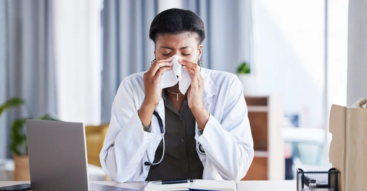 A physician in a white lab coat sits at a desk and blows her nose into a tissue, suggesting illness during flu season.