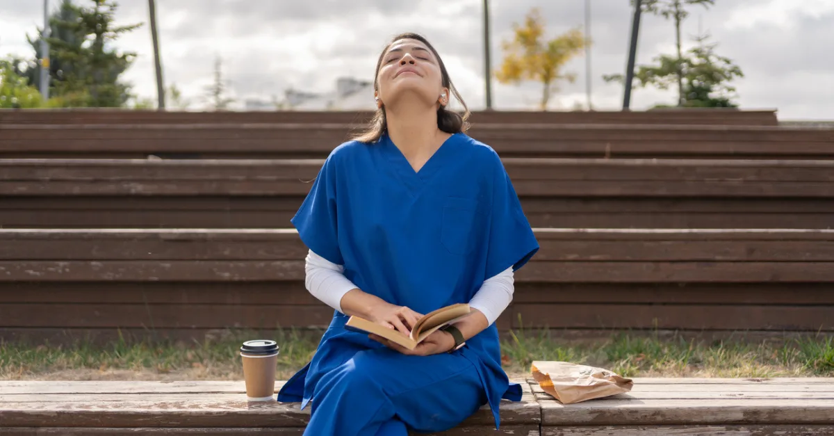 A healthcare worker in blue scrubs sits on an outdoor bench with eyes closed and head tilted back, taking a mindful break.
