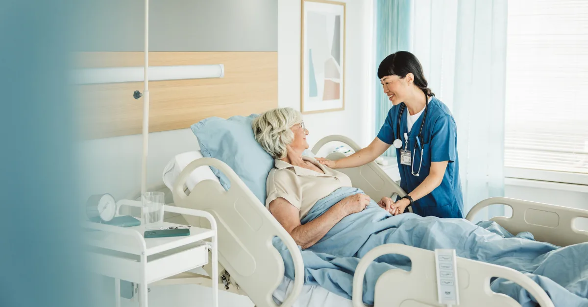 A nurse in blue scrubs with a stethoscope smiles while attending to a woman in a hospital bed.