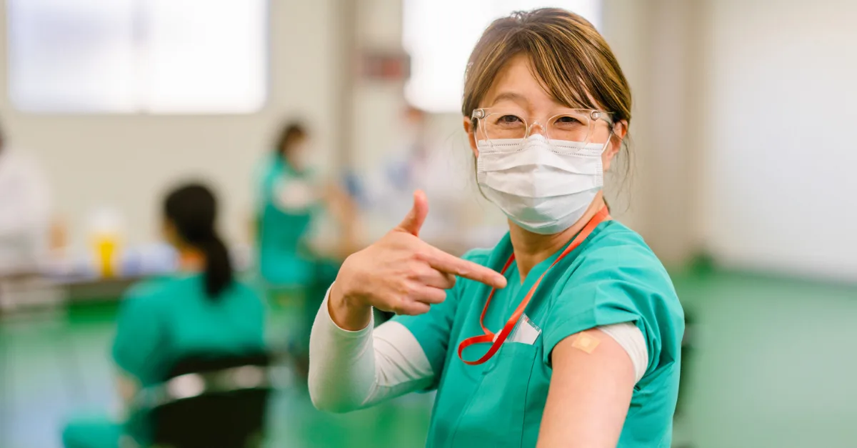 A healthcare worker in green scrubs, wearing a face mask and safety glasses, smiles while pointing to a bandage on their upper arm after receiving a vaccination.