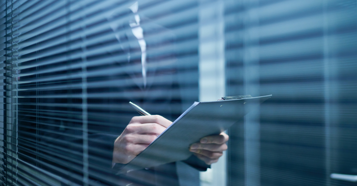 Silhouette of a professional in a suit writing on a clipboard, seen through office window blinds.