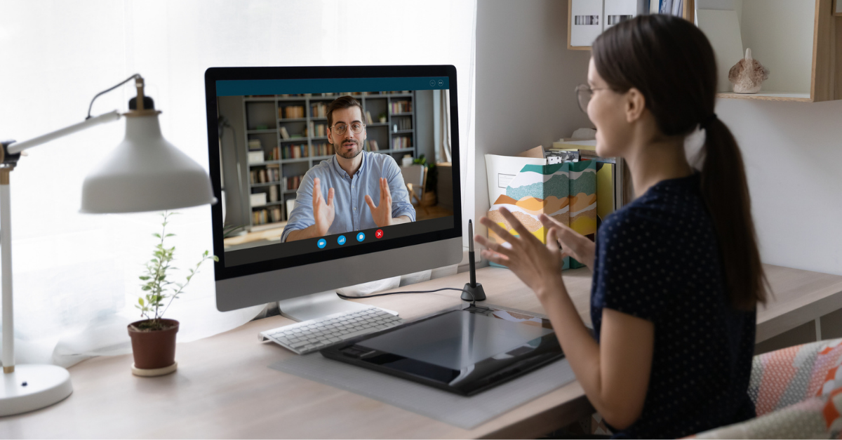 Woman at a home desk having a video call with a man, both gesturing while talking, in a bright office setting.